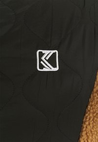 Čierny prešívaný materiál s diagonálnym stehom obsahuje biele vyšívané logo. Na jednej strane je prítomný kontrastný béžový textúrovaný materiál.