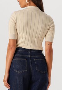 Beige kortærmet strikket sweater med lodrette ribber, med krave og subtile kontrasterende linjer. Sammen med mørkeblå højtaljede jeans.