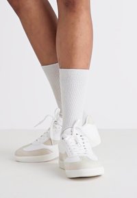 Baltie sneakers ar gaiši pelēkiem akcentiem, gluda ādas augšdaļa, teksturēta gumijas zole un baltas ribotas zeķes.
