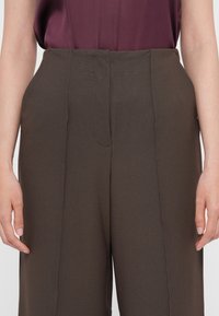 Pantaloni marroni su misura con una superficie liscia, vita alta e dettagli delle cuciture frontali, abbinati a un top senza maniche prugna.