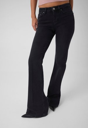 Femme portant un jean noir évasé taille haute et des chaussures noires à bout pointu, avec un haut court rose visible au-dessus de la taille.