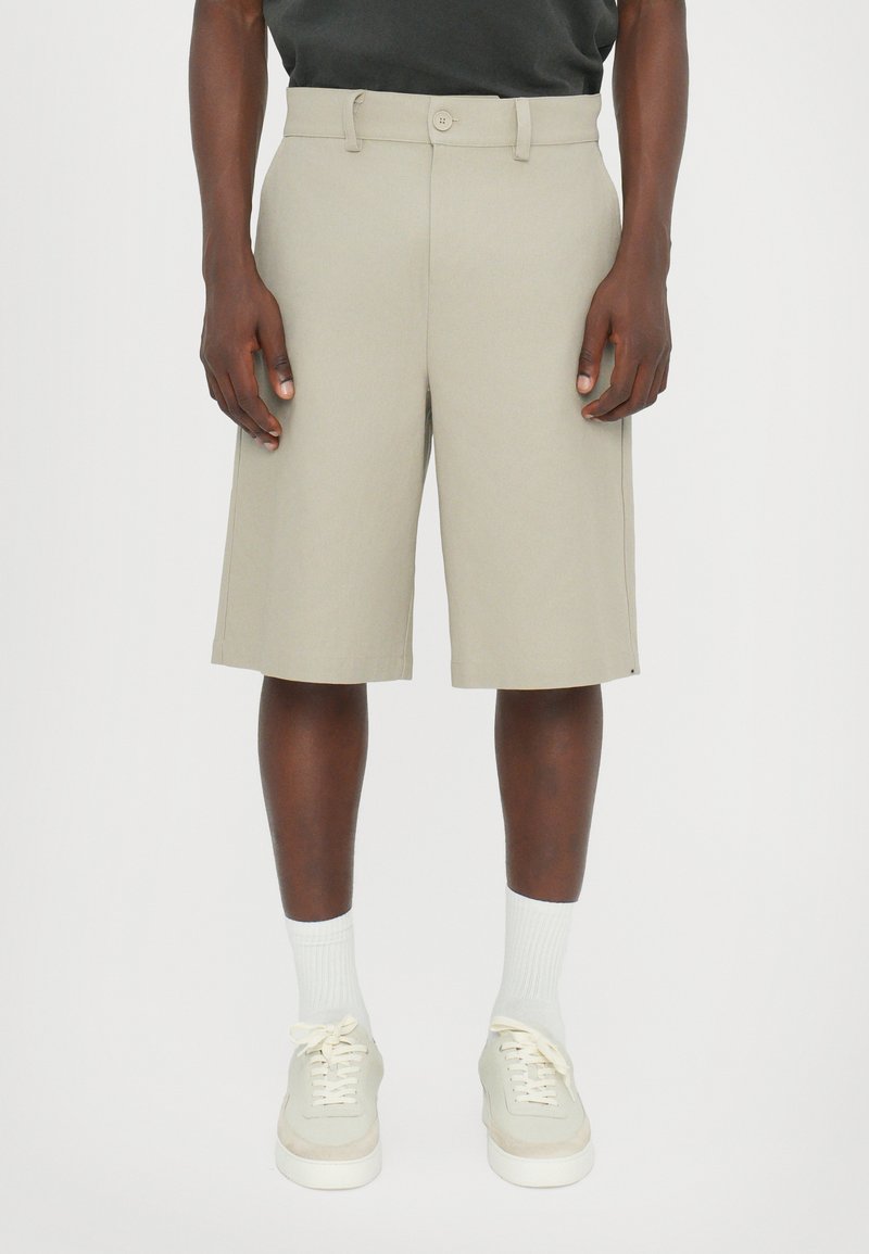 Shorts beige ajustés avec une coupe droite, passants de ceinture et une fermeture à un bouton, fabriqués à partir d'un tissu lisse. Portés avec des baskets et des chaussettes de couleur claire.