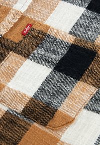 Tessuto flanella a quadri nei colori marrone, nero e crema con una superficie testurizzata; presenta un'etichetta di marca rossa e dettagli di cucitura visibili.