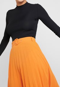 Maglietta nera a maniche lunghe abbinata a una gonna plissettata arancione con una cintura larga e una fibbia rettangolare, che presenta una texture liscia.