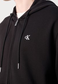 Gros plan sur une personne portant un sweat à capuche noir zippé avec le logo cK brodé en blanc sur la poitrine, des cordons noirs et un tirette de fermeture éclair argentée.