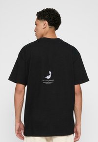 Sort oversized t-shirt med korte ærmer, prydet med et hvidt fjer grafik og tekst på bagsiden, lavet af bomuldsmateriale.