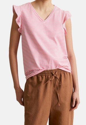 Persoon draagt een roze-wit gestreept mouwloos topje met gerimpelde schouders en bruine broeken met trekkoord, staat met ontspannen handen.