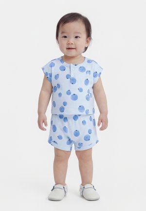 Kisgyermek előre nézve, világoskék felsőt és rövidnadrágot visel, kék gyümölcsmintákkal, valamint fehér cipőben, egy egyszerű háttér előtt.