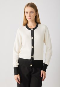 Cardigan tricoté noir et blanc avec des manches longues, des poignets côtelés et une fermeture à boutons avec des boutons métalliques. Texture douce, design ajusté.