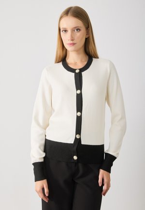 Valkoinen ja musta neulecardigan, jossa on pitkät hihat, ribattu rakenne ja metalliset napit. Pehmeä rakenne, istuva malli.