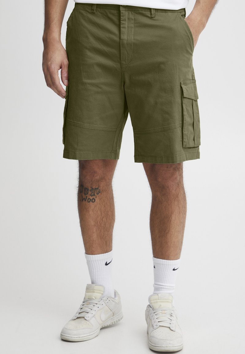 Shorts cargo verts olive en tissu à texture lisse. Présentent des poches latérales et une coupe regular, associés à des chaussettes de sport blanches et à des baskets.