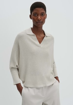 Frau mit kurzen Haaren, die einen hellbeigen, strukturierten Strickpullover mit Kragen und weiße Hose trägt, steht mit den Händen in den Taschen.