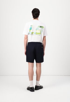 Homme portant un t-shirt graphique blanc, un short noir, des chaussettes blanches et des chaussures noires, debout face à un mur blanc uni.