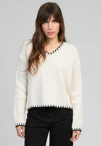 Pull en tricot crème avec un col en V, bord noir et texture côtelée. Coupe courte et carrée avec des détails en contraste au niveau de l'ourlet et des poignets.