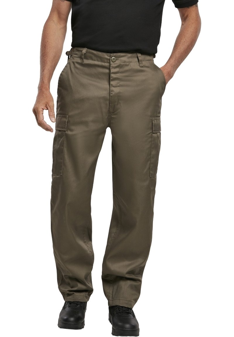 Homme portant un pantalon cargo vert olive, une ceinture noire, des chaussures noires et un t-shirt noir, debout avec une main dans la poche.
