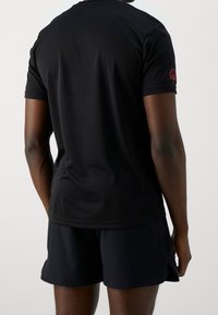 Camiseta atlética negra de manga corta con una textura suave, que presenta un pequeño logo rojo en el hombro derecho. Combinada con pantalones cortos oscuros.