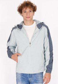 Giacca azzurro chiaro con un accento grigio scuro sulle spalle e sulle maniche. Caratterizzata da una chiusura con zip frontale e cappuccio. Tessuto leggero e liscia.
