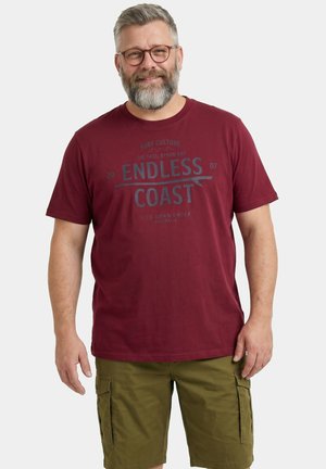 Erwachsener Mann mit Brille und Bart, der lächelt, trägt ein weinrotes "Endless Coast" T-Shirt und olivgrüne Cargo-Shorts, steht vor weißem Hintergrund.