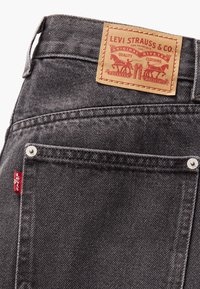Tumši pelēki denima džinsi ar redzamu ādas etiķeti, uz kuras rakstīts "Levi Strauss & Co.", un mazu sarkanu birku netālu no muguras kabatas.
