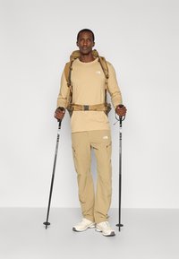 Μπεζ πουκάμισο μακριάς μανσέτας και παντελόνι καμφορντ, συνδυασμένα με σακίδιο πλάτης και μπαστούνια πεζοπορίας, με λογότυπο στο πουκάμισο και το παντελόνι.