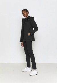 Veste imperméable noire avec capuche, fermeture éclair et poches latérales, assortie à un pantalon noir et des baskets blanches sur un fond clair.