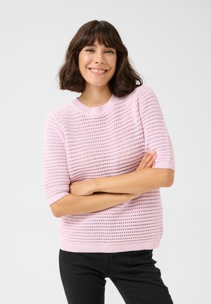 Pull en tricot rose clair avec un décolleté dégagé, des manches courtes et un motif ajouré. Associé à un pantalon noir.