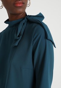 Teal blouse van gladde stof, met een hoge hals en een grote strikdetail bij de schouder, voorzien van een klein borstzakje.
