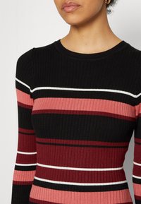 Femme portant un pull en maille côtelée avec des rayures horizontales en noir, bordeaux, rose et blanc, visible du cou à la taille.