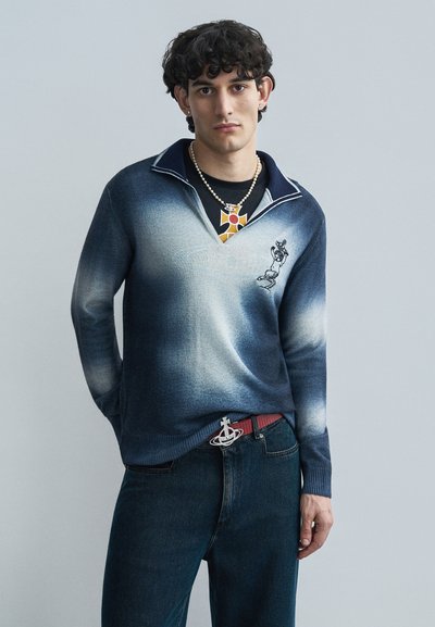 Μπλε πουλόβερ με διαβάθμιση στο πλεκτό και γιακά, με γραφικό στο στήθος, συνδυασμένο με σκουρόχρωμο τζιν και κόκκινη ζώνη με έμβλημα.