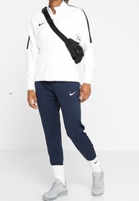 Weiße Zip-Jacke mit schwarzen Akzenten, marineblaue tapered Pants, graue Sneakers und eine schwarze Umhängetasche. Das Nike-Logo ist auf allen Artikeln sichtbar.