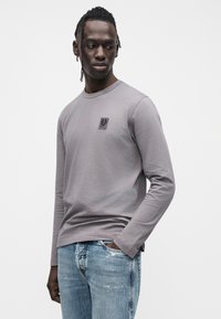 Pilkas ilgomis rankovėmis marškinėlis iš minkšto audinio, su mažu juodu logotipo ženkleliu ant krūtinės, derinamas su pašviesintais mėlynais džinsais.