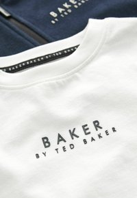 Camiseta de algodón blanca con un logo negro en relieve "BAKER by TED BAKER" en el pecho, sobre una prenda de forro polar azul marino oscuro.
