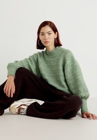 Pull en tricot vert avec un col montant et une coupe ample, associé à un pantalon large sombre et des baskets marron et blanches. Texture lisse.