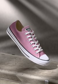 Pink canvas sneaker with white rubber sole, black accents, and white laces. Features a rounded toe and logo patch on the tongue.