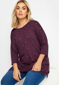 Lila tunika med trekvartslånga ärmar, prydd med spridda silverstjärnor. Tillverkad av mjukt tyg med en avslappnad passform och sidofickor.