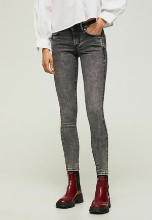 Person iført slidte sorte skinny jeans og skinnende røde ankelstøvler, stående imod en ensfarvet baggrund.