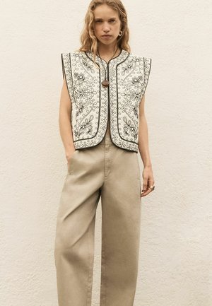 Mladá žena v čierno-bielej kvietkovanej vyšívanej veste, nad béžovými širokými nohavicami, stojaca pred štruktúrovanou svetlou stenou.