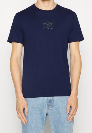 Osoba nosząca granatowy t-shirt z małym logo EA7 Emporio Armani na piersi oraz jasnoniebieskie dżinsy, stojąca na jednolitym tle.