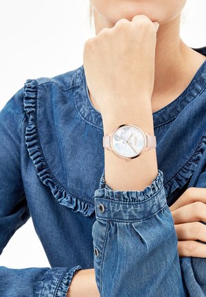 Runde Uhr mit Gehäuse aus Roségold, weißem Gummiband und floralem Zifferblatt, getragen zu einem Jeanshemd mit Rüschen.