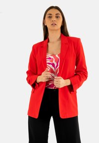 Blazer rojo con solapas de muesca y puños abotonados, combinado con una blusa rosa de estampado. Ubicado frente a un fondo neutro.