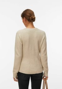 Femme aux cheveux bruns attachés en chignon, portant un pull beige, un jean noir, et tenant un sac à main en cuir beige sur un fond blanc uni.