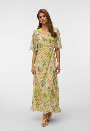 Femme en robe portefeuille jaune à fleurs avec des manches volantées et un ourlet froncé, portant des talons beiges à brides, debout devant un fond blanc uni.