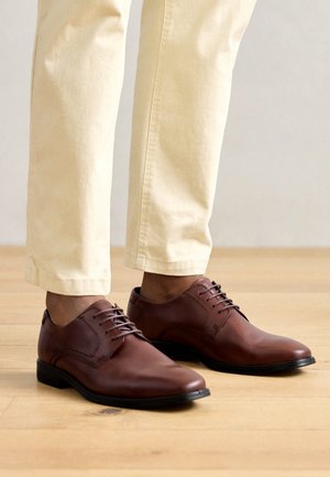 Chaussures habillées en cuir marron avec une texture lisse, un bout arrondi et un design à lacets. Assorties à un pantalon de couleur claire sur un sol en bois.