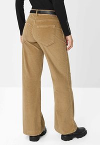 Pantaloni a gamba larga in velluto corduroy marrone con una superficie testurizzata, due tasche posteriori e una cintura nera. Stivaletti neri chunky sono visibili in basso.