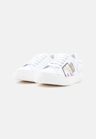 Άσπρα αθλητικά παπούτσια με κορδόνια, χοντρές σόλες και πολύχρωμο γραφικό αρκουδάκι στο πλάι, εμφανιζόμενα από μπροστινή γωνία σε λευκό φόντο.