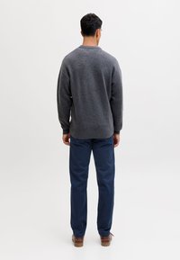 Mand iført mørkegrå sweater, marineblå bukser og brune sko, stående med ryggen mod et ensfarvet lyst baggrund.