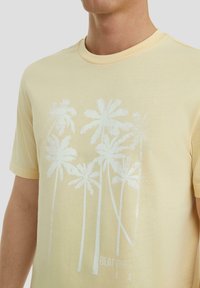 Lichtgele t-shirt met een witte palmboom grafiek en gedeeltelijk zichtbare tekst op de borst, gedragen door een persoon die is afgeknipt van de schouders naar beneden.