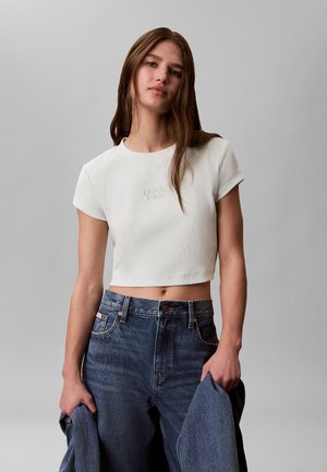 Ung kvinde iført en hvid cropped T-shirt og højtaljede blå jeans, der holder en denimjakke med en neutral grå baggrund.