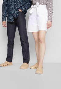 Två individer står bredvid varandra. Den ena har på sig mörka jeans och en mörk skjorta med bladmönster; den andra har vita shorts med en rosett och beige träskor.