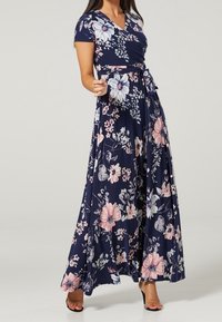 Woman wearing a navy blue floral wrap dress with a tie waist and short sleeves, paired with black high-heeled sandals.